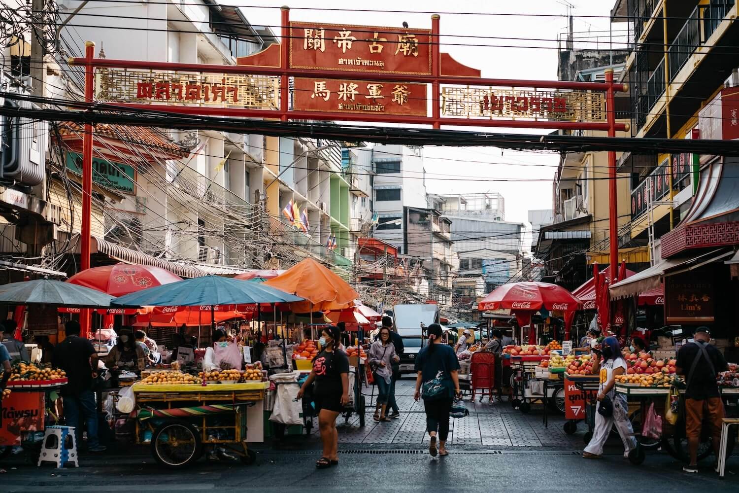 Bustling street market with fruit vendors beneath a red Chinese archway in Chinatown, Bangkok. 
