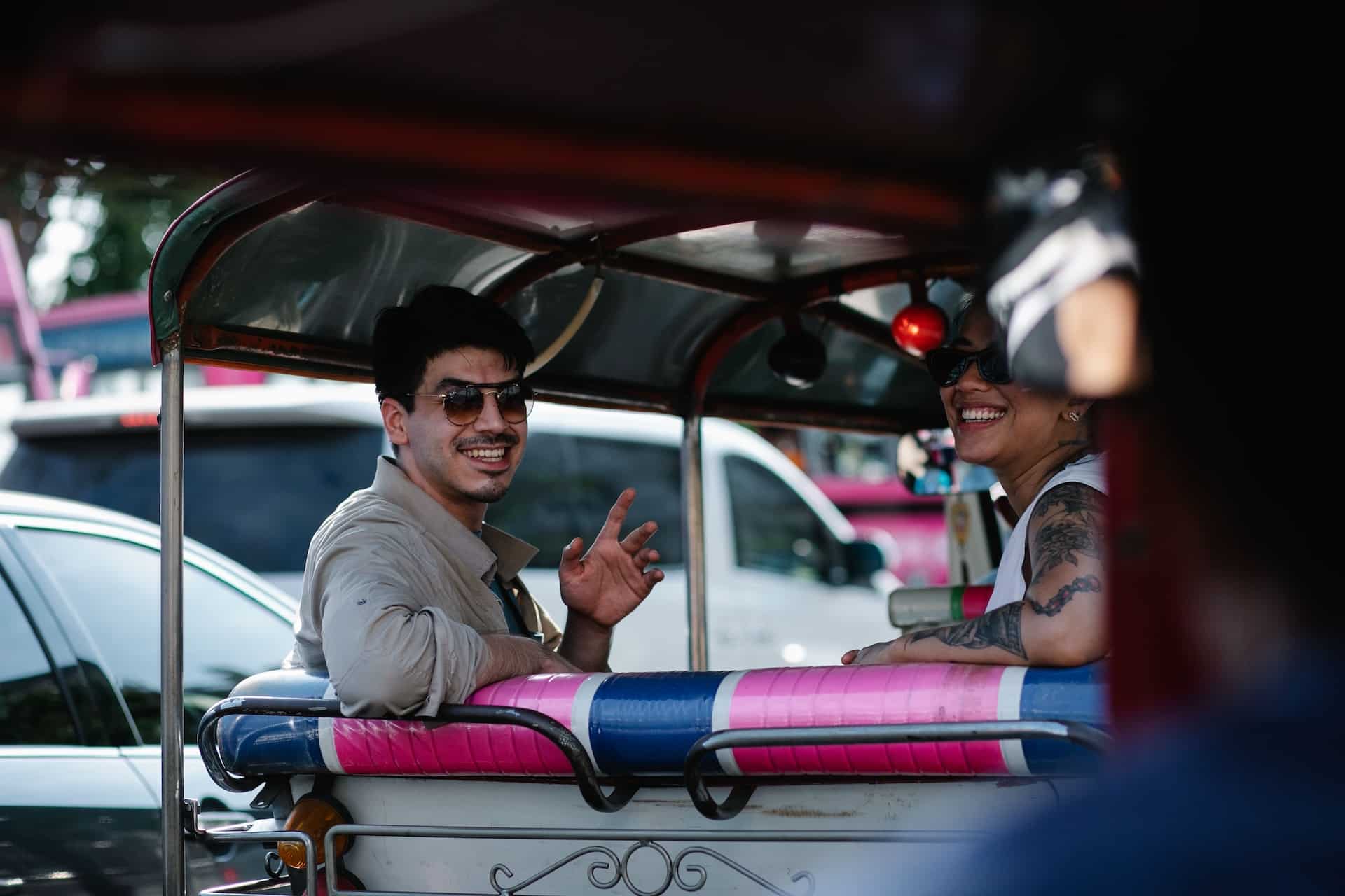 A smiling man and woman ride together in the back of a tuk-tuk while exploring Bangkok.