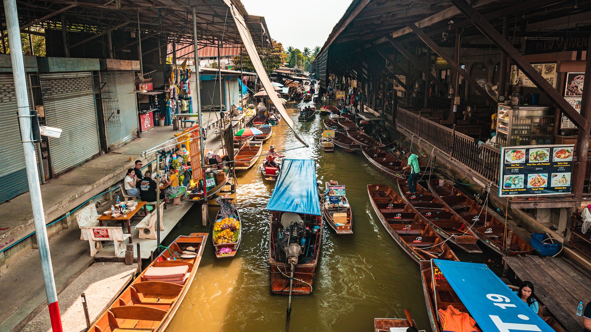 High-angle view of wooden longtail boats navigating a canal flanked by covered shopfronts at a Thai floating market.